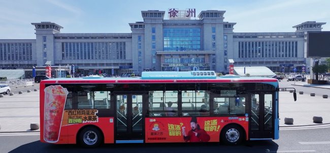 图为徐州东站站前广场，涂装着蜜城之恋西红柿鸡蛋汤的本地茶饮品牌广告公交车行驶在站前主干道上